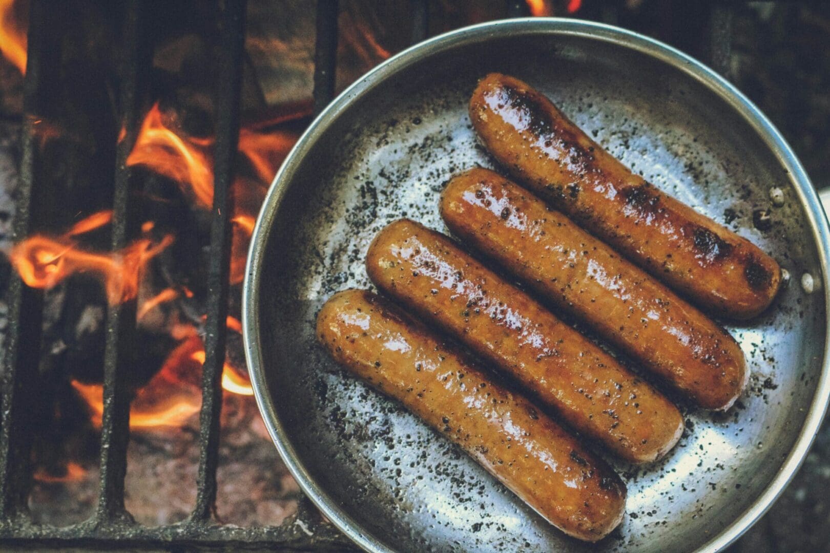 A pan of hot dogs cooking over an open fire.